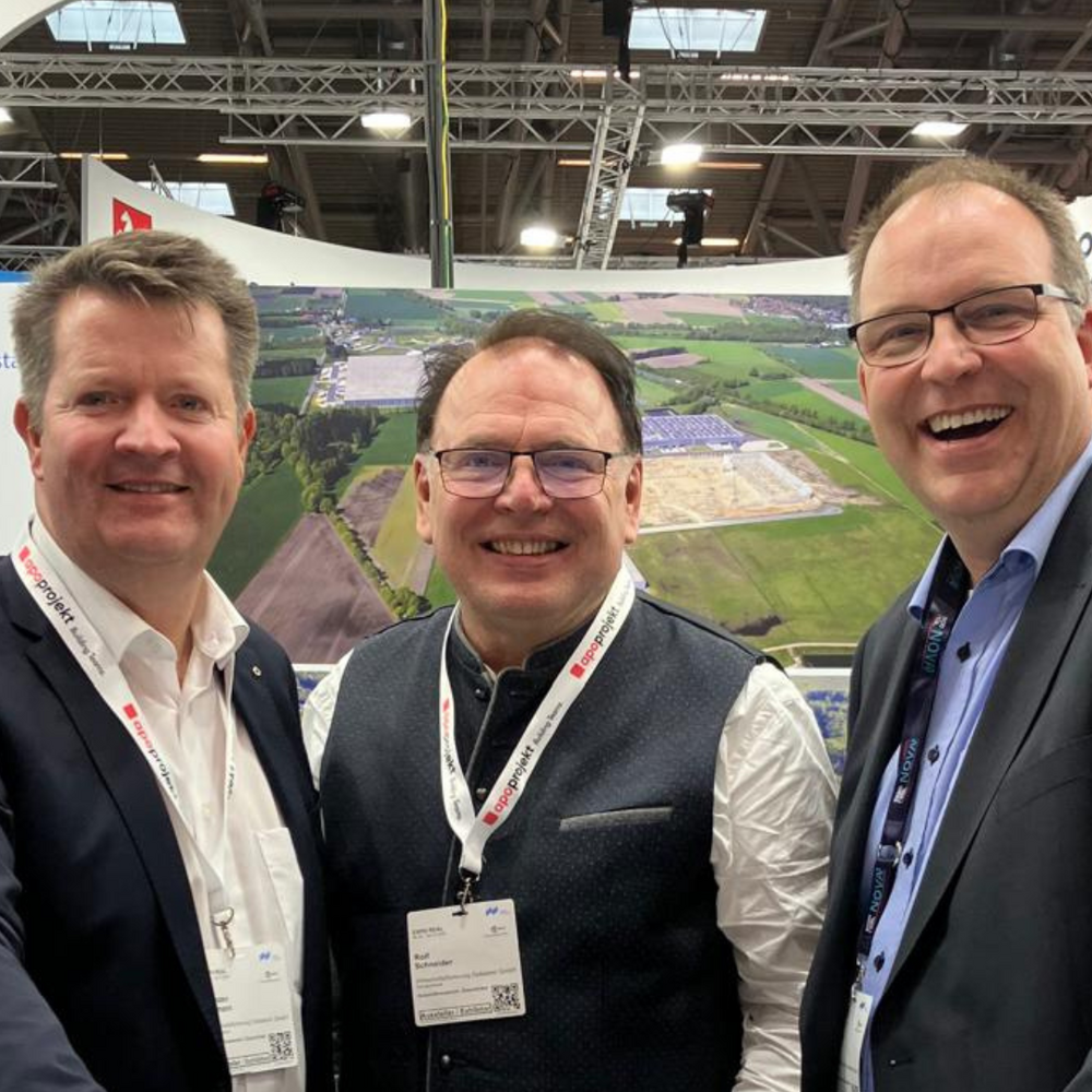 | Ⓒ Deltaland Auf dem Bild sind drei Männer zu sehen, die lächelnd vor einem Tisch stehen und sich in einer Messeumgebung befinden. Im Hintergrund werden Informationen über die Region Deltaland angezeigt.