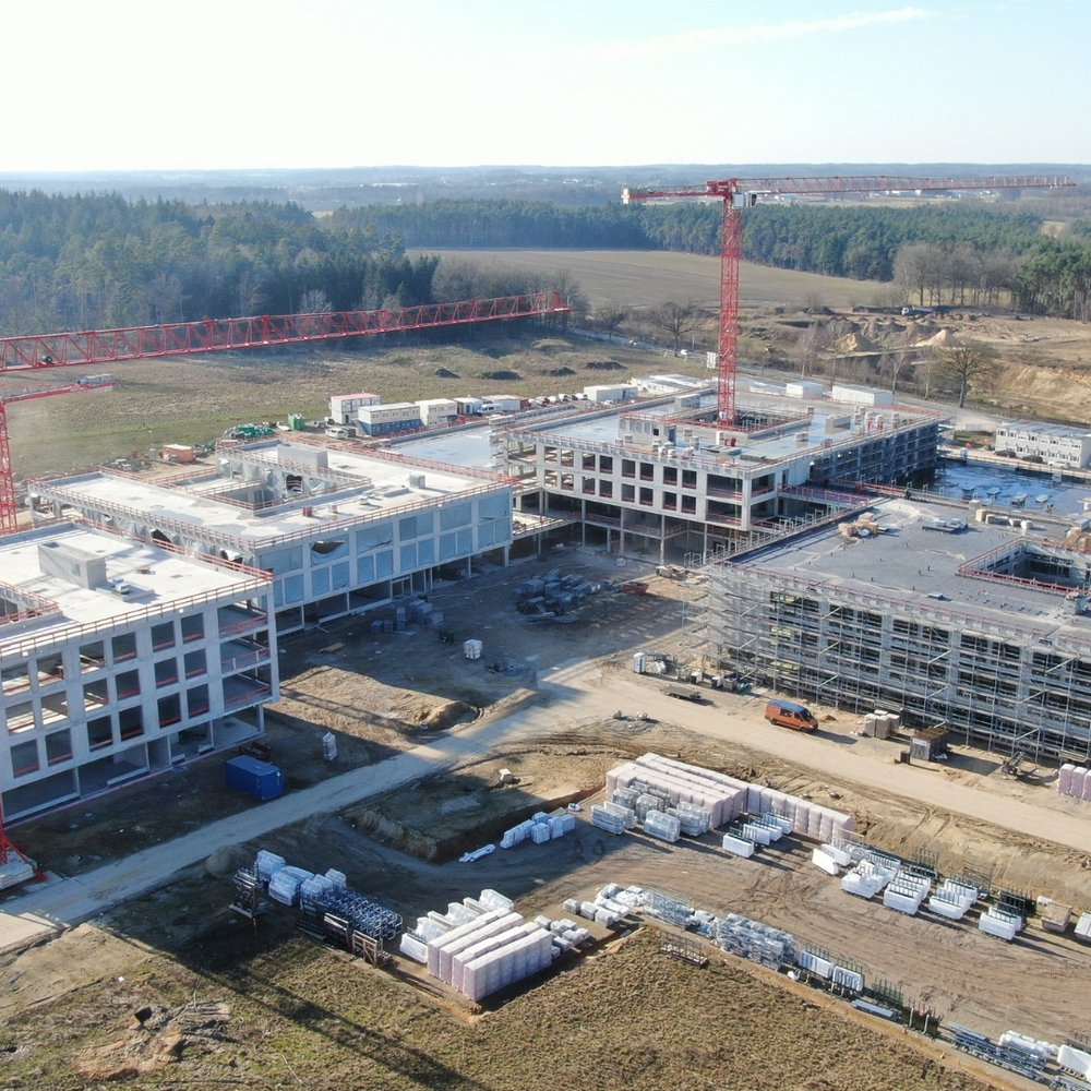 | Ⓒ Heidekreis Klinikum Das Luftbild zeigt eine große Baustelle mit mehreren mehrstöckigen Gebäuden im Rohbau, die von zwei markanten roten Liebherr-Turmdrehkranen überragt werden. Das weitläufige Gelände ist umgeben von Waldgebieten und offenen Feldflächen, während auf dem Baufeld zahlreiche Baumaterialien und Baufahrzeuge zu erkennen sind.