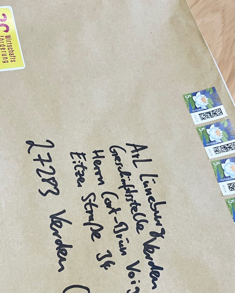 Das Bild zeigt einen braunen Briefumschlag mit handschriftlicher Adresse. Er ist mit mehreren Briefmarken frankiert. Oben links befindet sich ein gelber Aufkleber mit einem Jubiläumshinweis. Der Umschlag liegt auf einer Holzoberfläche.