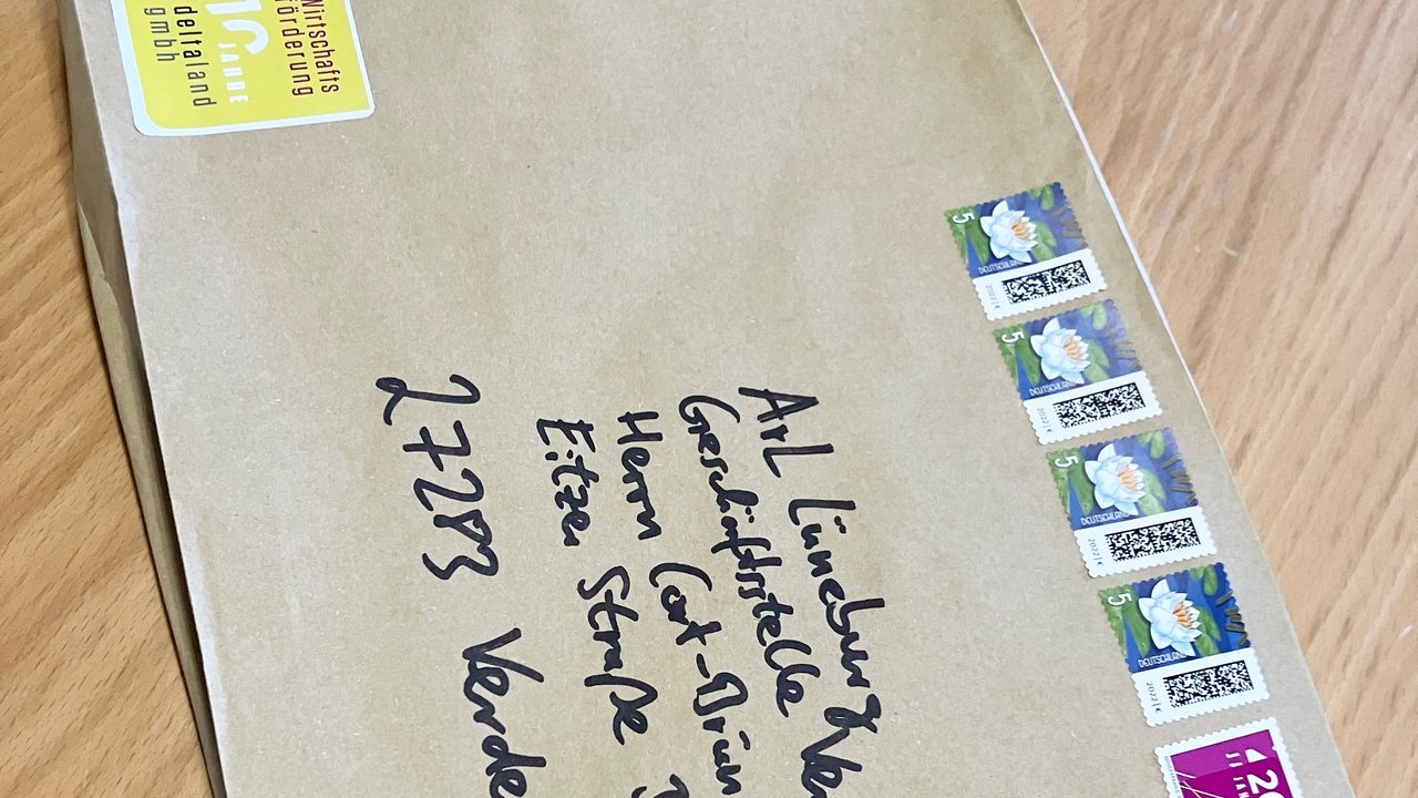 Das Bild zeigt einen braunen Briefumschlag mit handschriftlicher Adresse. Er ist mit mehreren Briefmarken frankiert. Oben links befindet sich ein gelber Aufkleber mit einem Jubiläumshinweis. Der Umschlag liegt auf einer Holzoberfläche.