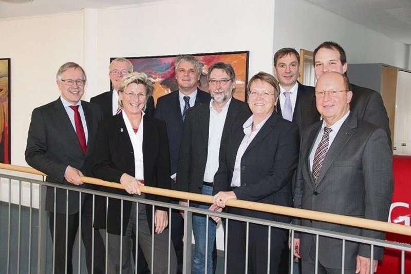 Das Bild zeigt eine Gruppe von Politikern und Vertretern einer Behörde oder Organisation, die gemeinsam für ein Foto posieren. Sie stehen in einem Bürogebäude vor einem Gemälde an einer Wand. Die Personen tragen überwiegend formelle Kleidung wie Anzüge und Kostüme, was auf einen offiziellen Anlass hindeutet. Insgesamt vermittelt das Bild einen Eindruck von Professionalität und Zusammenarbeit innerhalb der Gruppe.