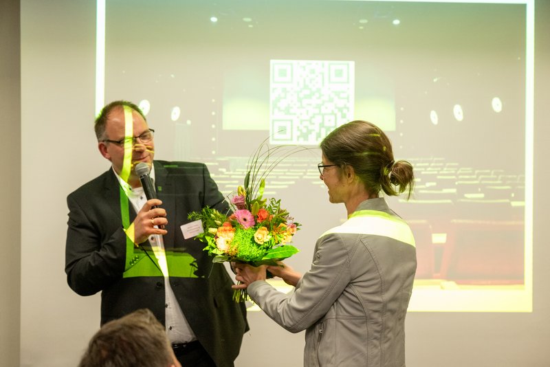 Das Bild zeigt einen Mann, der mit einem Mikrofon in der Hand eine Frau anspricht, während sie einen Blumenstrauß entgegennimmt. 