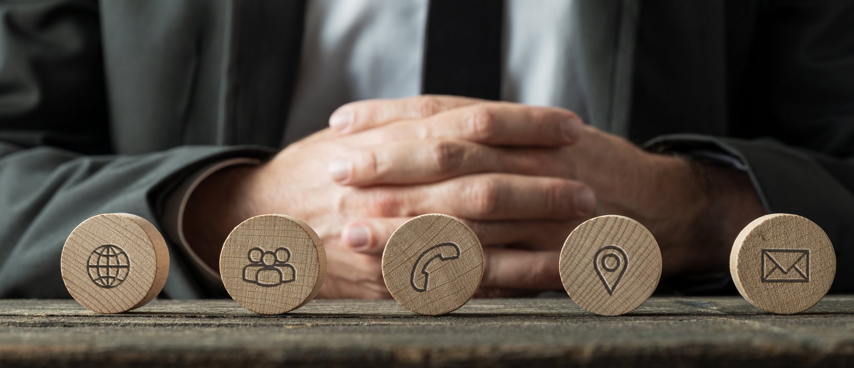  Das Bild zeigt eine Person in einem Anzug, die hinter einer Reihe von fünf Holzscheiben sitzt. Auf den Holzscheiben sind Symbole eingraviert, die verschiedene Kommunikationsmethoden darstellen. Von links nach rechts sind es: ein Globus für das Internet, eine Gruppe von Personen für Teamarbeit, ein Telefonhörer, eine Standortmarkierung und ein Briefumschlag für E-Mail. Die Person hat die Hände vor sich verschränkt und trägt einen dunklen Anzug.