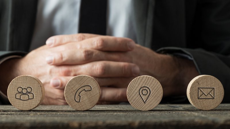 Das Bild zeigt eine Person in einem Anzug, die hinter einer Reihe von fünf Holzscheiben sitzt. Auf den Holzscheiben sind Symbole eingraviert, die verschiedene Kommunikationsmethoden darstellen. Von links nach rechts sind es: ein Globus für das Internet, eine Gruppe von Personen für Teamarbeit, ein Telefonhörer, eine Standortmarkierung und ein Briefumschlag für E-Mail. Die Person hat die Hände vor sich verschränkt und trägt einen dunklen Anzug.