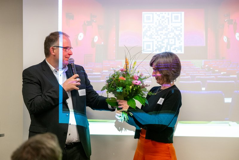 Das Bild zeigt einen Mann, der mit einem Mikrofon in der Hand eine Frau anspricht, während sie einen Blumenstrauß entgegennimmt. Im Hintergrund ist ein Kinosaal zu sehen, der mit einer Präsentation oder einem QR-Code beleuchtet ist.