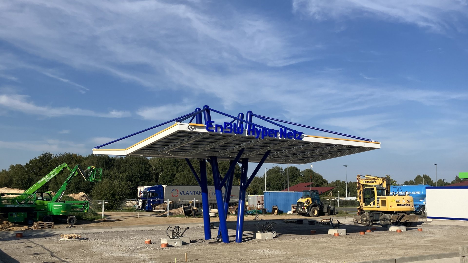 Das Bild zeigt eine Baustelle, auf der eine moderne „EnBW HyperNetz“-Ladestation errichtet wird, unterstützt von blauen Säulen unter einem futuristischen Dach. Rund um die Station sind Baugeräte wie ein Kran und ein Bagger sowie ein LKW im Hintergrund zu sehen.