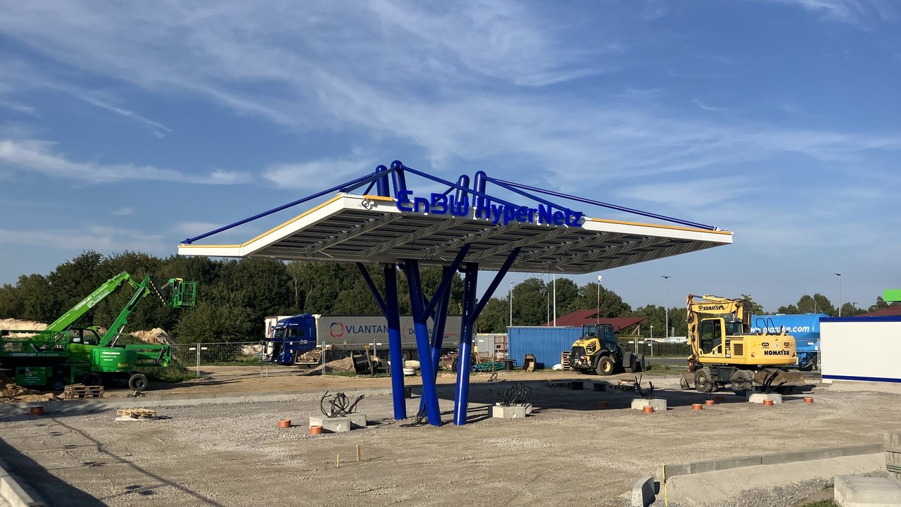 Das Bild zeigt eine Baustelle, auf der eine moderne „EnBW HyperNetz“-Ladestation errichtet wird, unterstützt von blauen Säulen unter einem futuristischen Dach. Rund um die Station sind Baugeräte wie ein Kran und ein Bagger sowie ein LKW im Hintergrund zu sehen.