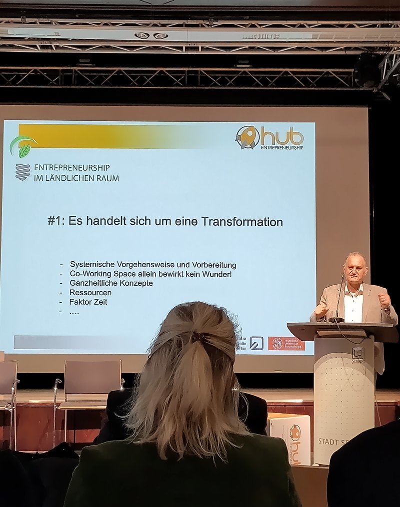 Das Bild zeigt einen Redner auf einer Bühne, der vor einem Publikum einen Vortrag hält. Auf der Leinwand hinter ihm steht der Titel "#1: Es handelt sich um eine Transformation" mit weiteren Stichpunkten zur systemischen Vorgehensweise, ganzheitlichen Konzepten, Ressourcen und dem Faktor Zeit.