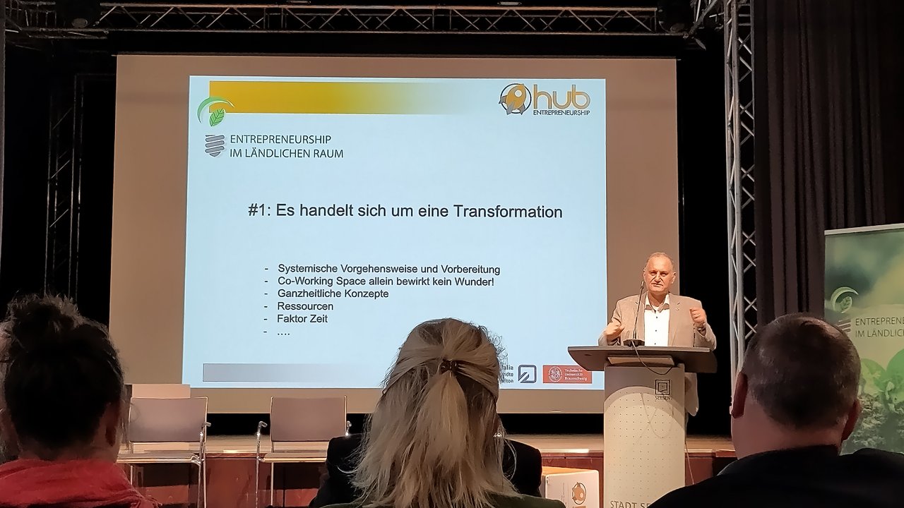 Das Bild zeigt einen Redner auf einer Bühne, der vor einem Publikum einen Vortrag hält. Auf der Leinwand hinter ihm steht der Titel "#1: Es handelt sich um eine Transformation" mit weiteren Stichpunkten zur systemischen Vorgehensweise, ganzheitlichen Konzepten, Ressourcen und dem Faktor Zeit.