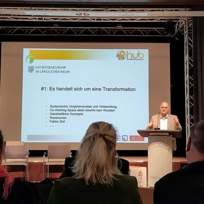 Das Bild zeigt einen Redner auf einer Bühne, der vor einem Publikum einen Vortrag hält. Auf der Leinwand hinter ihm steht der Titel "#1: Es handelt sich um eine Transformation" mit weiteren Stichpunkten zur systemischen Vorgehensweise, ganzheitlichen Konzepten, Ressourcen und dem Faktor Zeit.