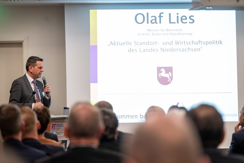 Das Bild zeigt einen Mann, der an einem Rednerpult steht und in ein Mikrofon spricht, während er zu einem sitzenden Publikum spricht. Im Hintergrund ist eine Projektion zu sehen, auf der „Olaf Lies“ und der Titel „Aktuelle Standort- und Wirtschaftspolitik des Landes Niedersachsen“ mit dem niedersächsischen Wappen angezeigt werden.