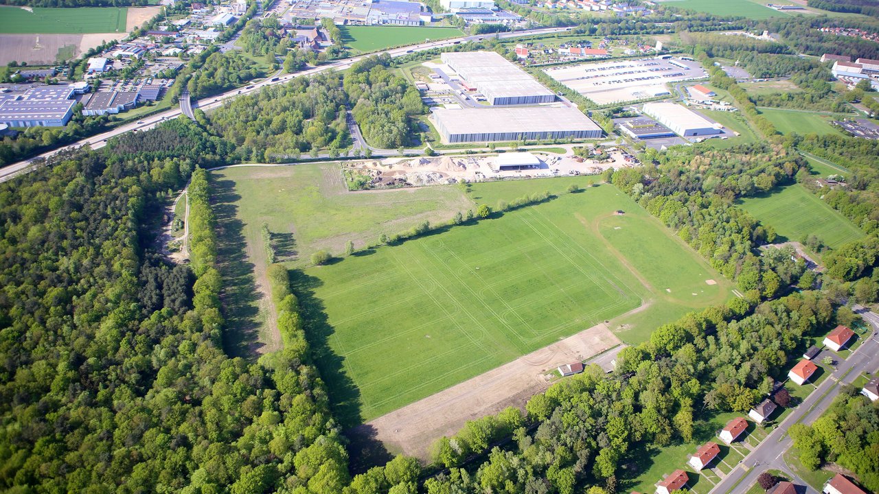  Das Bild zeigt eine weitläufige, grüne Landschaft mit Feldern, Wäldern und vereinzelten Gebäuden, darunter große Industriehallen im Vordergrund. Eine Autobahn verläuft durch das Bild, neben der sich eine Brücke befindet, die zu einem kleinen, von Bäumen gesäumten Weg führt.