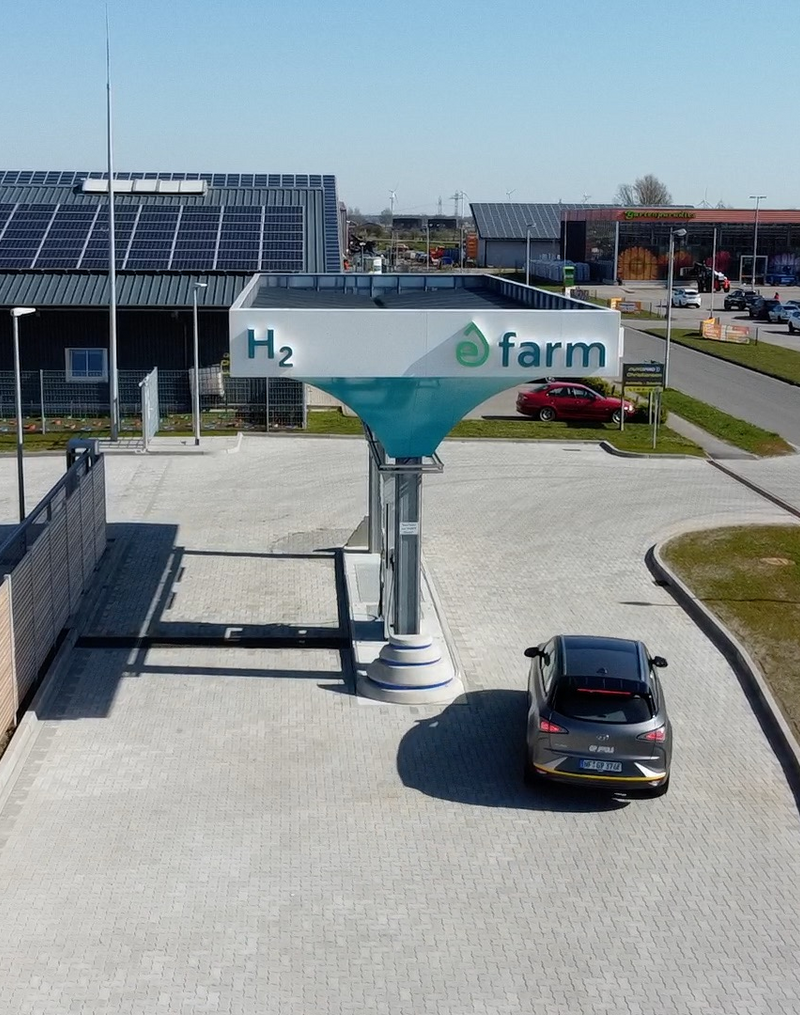 Das Bild zeigt eine moderne Wasserstofftankstelle namens "H2 farm", die an einer größeren Autohandelseinrichtung angesiedelt ist. Das Gebäude ist mit einer großflächigen Photovoltaikanlage ausgestattet, was auf eine nachhaltige Energieversorgung der Tankstelle hindeutet. Mehrere Autos sind an der Tankstelle zu sehen, was die Nutzung der Anlage durch Fahrer von Wasserstofffahrzeugen belegt.