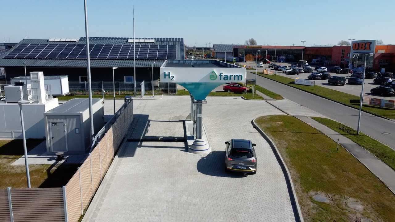 Das Bild zeigt eine moderne Wasserstofftankstelle namens "H2 farm", die an einer größeren Autohandelseinrichtung angesiedelt ist. Das Gebäude ist mit einer großflächigen Photovoltaikanlage ausgestattet, was auf eine nachhaltige Energieversorgung der Tankstelle hindeutet. Mehrere Autos sind an der Tankstelle zu sehen, was die Nutzung der Anlage durch Fahrer von Wasserstofffahrzeugen belegt.