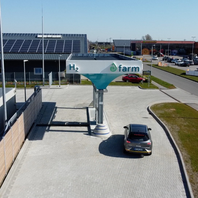 Das Bild zeigt eine moderne Wasserstofftankstelle namens "H2 farm", die an einer größeren Autohandelseinrichtung angesiedelt ist. Das Gebäude ist mit einer großflächigen Photovoltaikanlage ausgestattet, was auf eine nachhaltige Energieversorgung der Tankstelle hindeutet. Mehrere Autos sind an der Tankstelle zu sehen, was die Nutzung der Anlage durch Fahrer von Wasserstofffahrzeugen belegt.