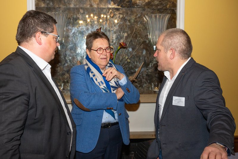 Das Bild zeigt drei Personen in einem angeregten Gespräch, wobei die Frau in der Mitte einen blauen Blazer und einen gemusterten Schal trägt. Im Hintergrund ist ein dekorativer Spiegel mit Blumenarrangement und Glasvasen zu sehen, was auf eine elegante Umgebung hindeutet.