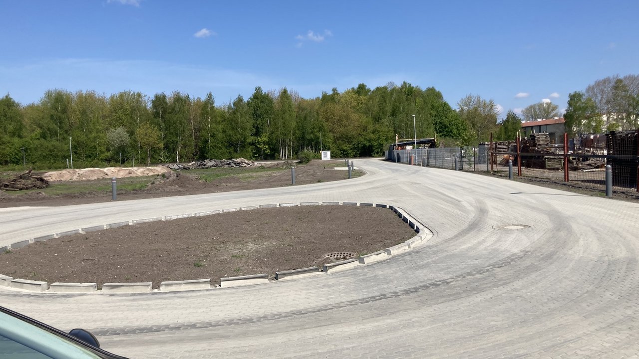  Das Bild zeigt eine neu gepflasterte Straße, die in eine Kurve mündet und von einem kreisförmigen Bereich mit unbepflanzter Erde umgeben ist. Im Hintergrund sind Bäume und einige Industriegebäude zu sehen, die den Straßenrand säumen.