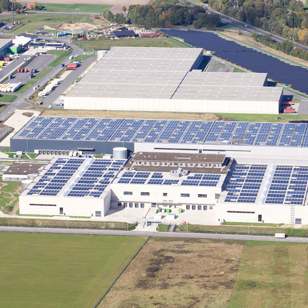 | Ⓒ Deltaland Das Bild zeigt eine Luftaufnahme einer großen Halle, die mit Solarzellen auf dem Dach ausgestattet ist, umgeben von weitläufigen Grünflächen und weiteren Lagerhallen. Die Landschaft im Hintergrund ist von Bäumen und Feldern geprägt.