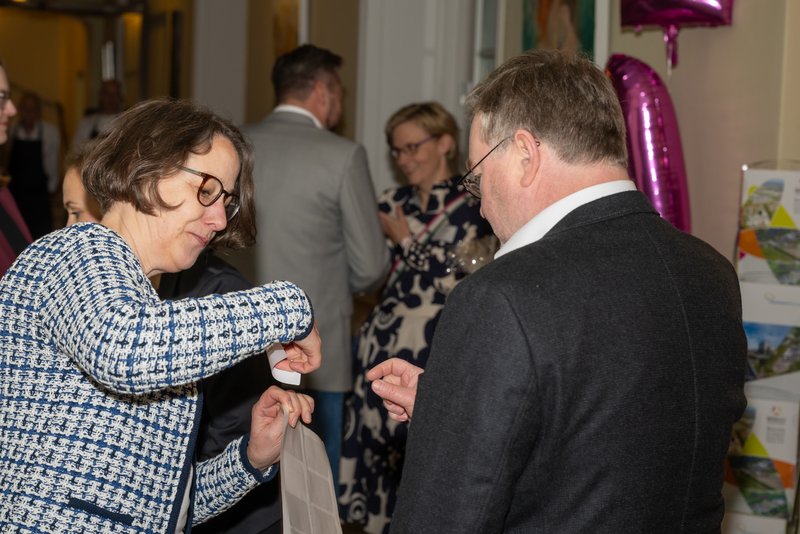 Auf dem Bild sind zwei Personen in einem geschäftlichen Umfeld zu sehen, die miteinander interagieren. Eine Frau mit Brille übergibt einem Mann ein Namensschild, während im Hintergrund weitere Gäste anwesend sind.