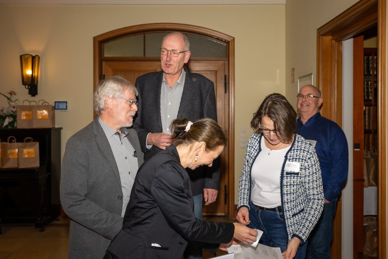 Das Bild zeigt eine Gruppe von vier Personen in einem Innenraum, die an einem Tisch beschäftigt sind. Zwei Männer stehen im Hintergrund, während eine Frau und ein Mann im Vordergrund Namensschilder verteilen und miteinander sprechen.