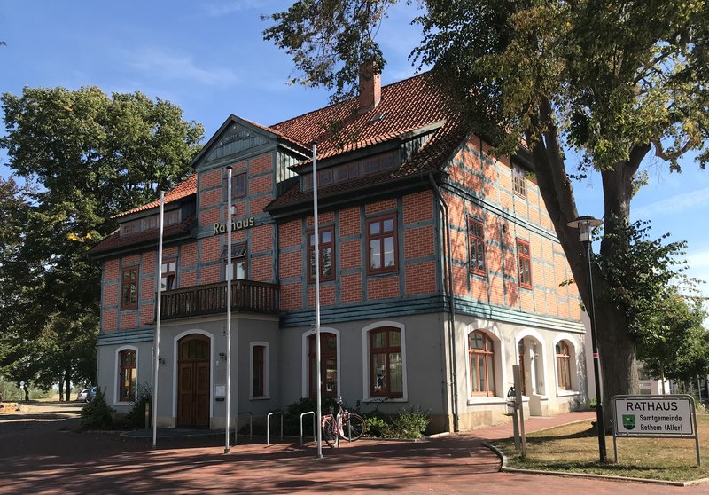 Das Bild zeigt ein historisches, rot-braunes Rathaus-Gebäude mit charakteristischer Architektur, das inmitten einer begrünten Umgebung steht. Der Fahrradständer und die Straßenschilder vor dem Gebäude deuten darauf hin, dass es sich um einen öffentlichen Verwaltungssitz handelt.