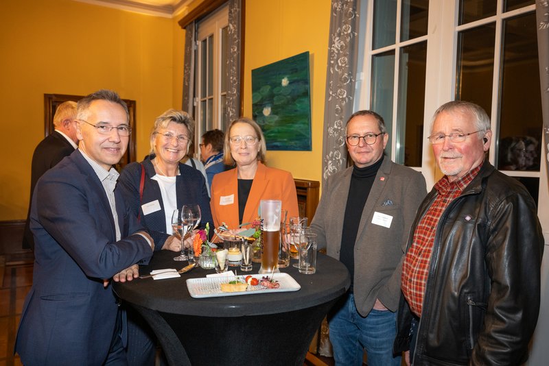 Das Bild zeigt fünf Personen, die an einem Stehtisch stehen und lächeln, während sie Getränke und Snacks vor sich haben. Die Umgebung wirkt festlich und einladend, was auf eine gesellige Veranstaltung hindeutet.