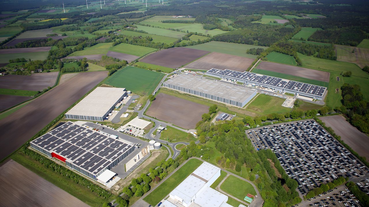  Das Bild zeigt eine große Industrieanlage mit mehreren Gebäuden und Lagerhallen, umgeben von einer ländlichen Landschaft mit Feldern, Wäldern und Windrädern. Es ist eine Luftaufnahme, die einen Überblick über das Industrieareal und seine Infrastruktur mit Parkplätzen, Straßen und Grünflächen gibt.