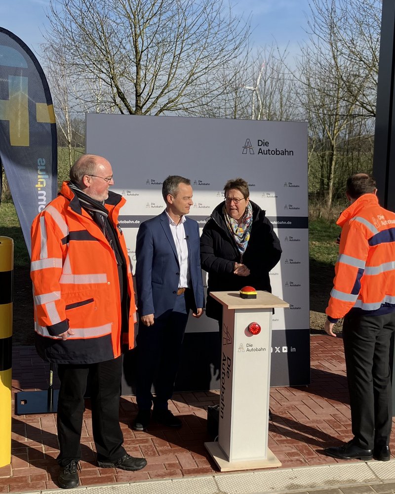 Das Bild zeigt eine Gruppe von vier Personen, die an einer modernen Ladestation stehen. Zwei Personen tragen orangefarbene Warnwesten, während die anderen beiden Personen formelle Kleidung tragen und mit ihnen sprechen, möglicherweise im Rahmen einer Einweihung. 