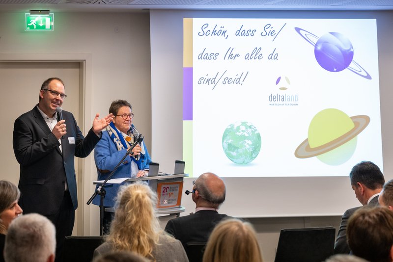 Auf dem Bild stehen ein Mann und eine Frau an einem Stehpult, beide halten ein Mikrofon hält und der Mann spricht. Vor ihnen sitzt ein Publikum, und das Rednerpult trägt ein Schild mit der Aufschrift „20 Jahre Wirtschaftsförderung DeltaLland GmbH“. Im Hintergrund ist eine Projektion zu sehen.