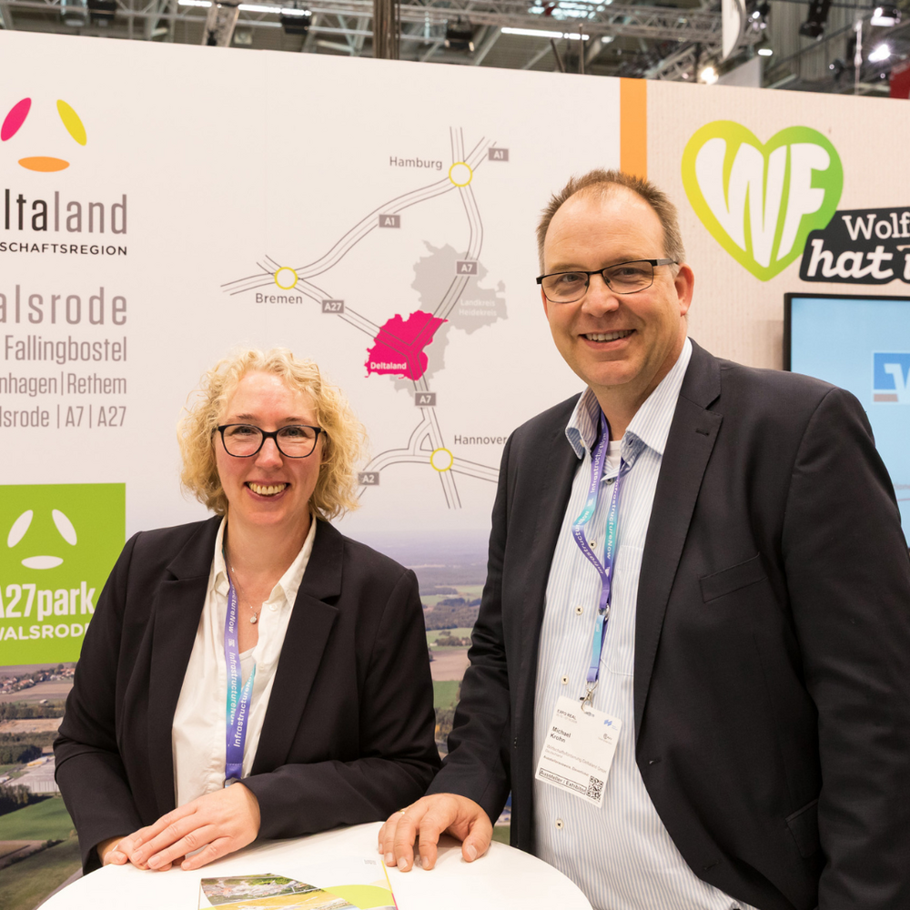 | Ⓒ Kalle Singer Eine Frau mit lockigen blonden Haaren und ein Mann mit Brille stehen lächelnd an einem Stehtisch am Messestand der Deltaland Wirtschaftsregion. Im Hintergrund sind das Deltaland-Logo, eine Anfahrtskarte und der A27park Walsrode zu sehen.